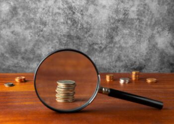 Imagem que mostra moedas sendo observadas através de uma lupa sobre uma mesa de madeira, representando a verificação de autenticidade de moedas.