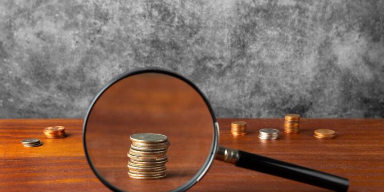 Imagem que mostra moedas sendo observadas através de uma lupa sobre uma mesa de madeira, representando a verificação de autenticidade de moedas.
