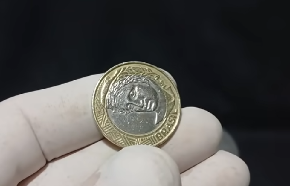 Moeda de 1 real com defeito de reverso horizontal sendo segurada por luva